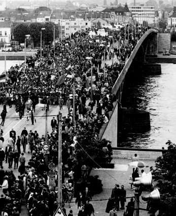 Mai 1968, Bonn: Demonstration von 70.000 Menschen gegen die Notstandsgesetze. Sie wissen, dass Demonstrationen nicht ausreichen, und fordern: die Arbeiter müssen in den Generalstreik treten! Der DGB-Vorstand aber lehnt dieses Kampfmittel grundsätzlich ab und tritt damit die zu dieser Zeit neu erwachende Kampfkraft der Arbeiter mit Füßen.